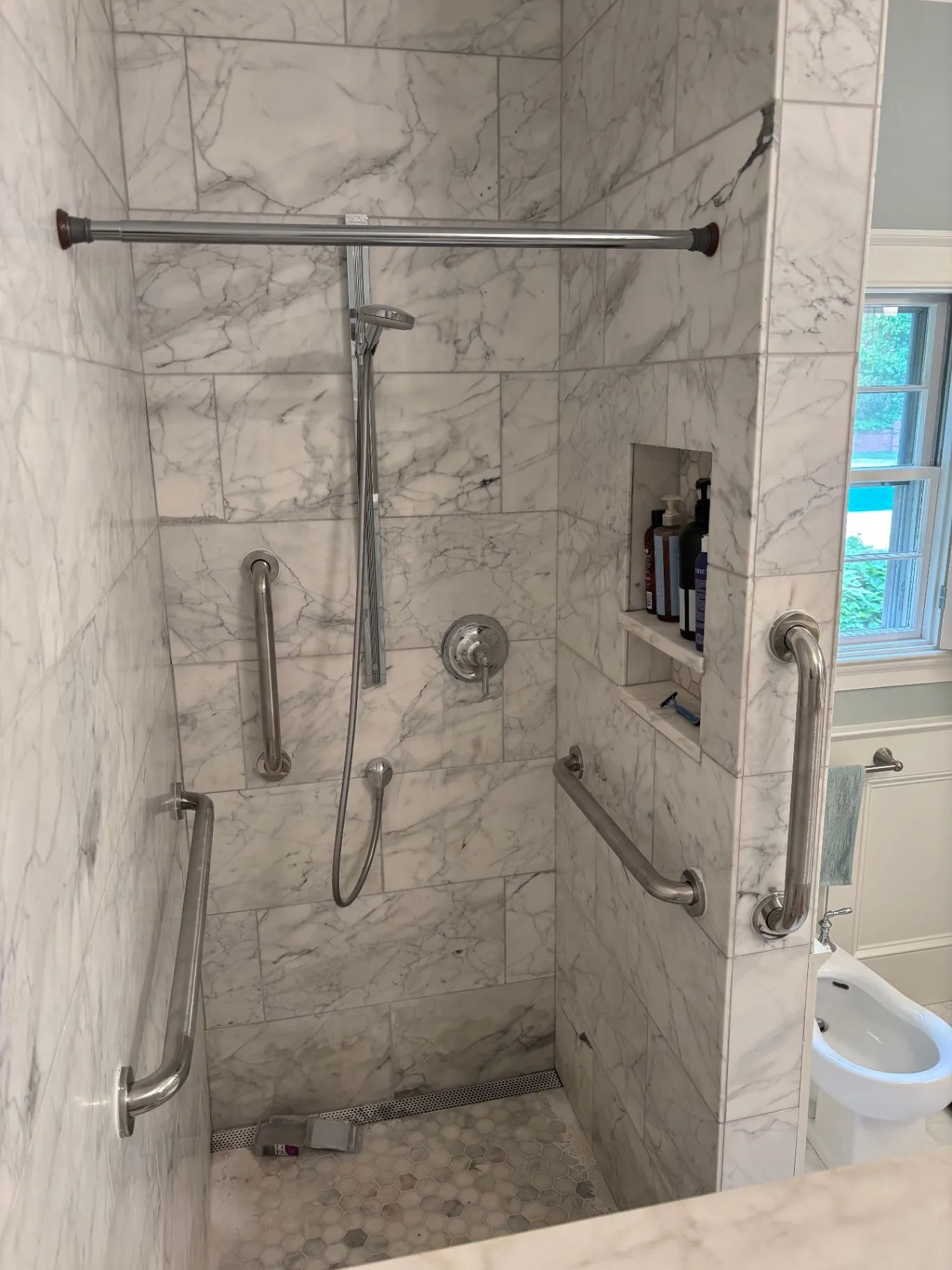 Marble-look tiled shower with stainless grab bars, a handheld shower, and a built-in shelf.